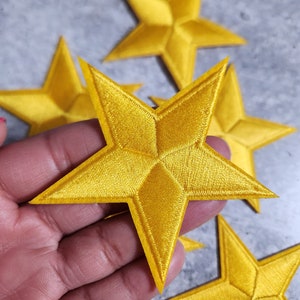 Peut inclure: Cinq patchs en tissu en forme d'étoile dorée. Les étoiles sont en 3D et ont une texture en relief. Les étoiles sont disposées en demi-cercle avec une étoile au centre.