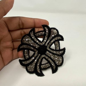 May include: A black and silver embroidered patch with a circular design. The center of the patch is black and the outer edges are silver.