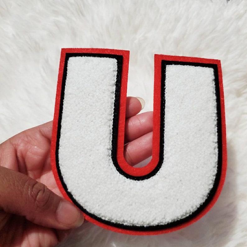 Puede incluir: Letra U de fieltro blanco con contorno negro y borde rojo. La letra es gruesa y tiene un efecto 3D.