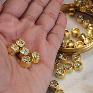 Peut inclure: Gros plan sur des ornements cylindriques dorés avec des centres en cristal clair et étincelant. Certains sont tenus dans une main, d'autres sont dispersés sur une surface et dans un petit récipient. Ces éléments décoratifs sont probablement destinés à l'artisanat ou à la bijouterie.