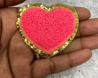 Puede incluir: Un parche en forma de corazón rosa con un borde de purpurina dorada. El corazón es peludo y el borde es brillante.