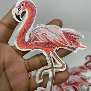May include: Pink flamingo embroidered iron-on patch. The flamingo is standing on one leg with its wings spread out. The patch is made of a white fabric with pink and brown embroidery.