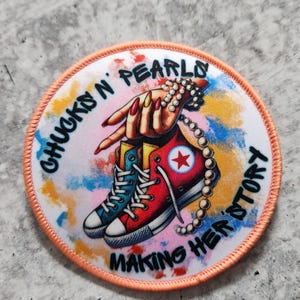 May include: A round, peach-coloured patch with a white background and a colourful design. The design features a hand holding a string of pearls and a red and blue sneaker with a white star. The text "Chucks 'n Pearls Making Her Story" is written around the edge of the patch.