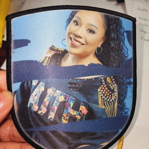 Puede incluir: Un parche en forma de escudo con una mujer sonriente y la palabra "KITH" en letras florales. El parche tiene un fondo azul y un borde negro. Un artículo decorativo.