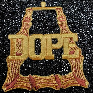 May include: A gold embroidered patch with the word "DOPE" in a stylized font. The patch has a red outline and is surrounded by a black background.