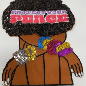 May include: A brown fabric patch with a black afro and the words "Protect Your Peace" in pink and white. The afro is covered in black sequins. The patch has a rainbow of sequins on the shoulder and a silver sequin bracelet on the arm.
