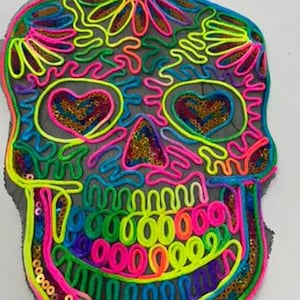 May include: A colorful, embroidered skull patch with sequins. The skull is outlined in neon pink, green, blue, and yellow thread. The eyes are outlined in pink and have sequins in the shape of hearts.