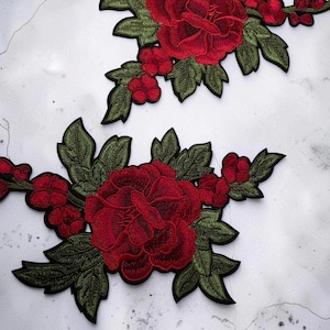 Op de afbeelding: Twee geborduurde patches met grote, donkerrode bloemen met gelaagde bloemblaadjes en donkergroene bladeren. De bloemmotieven zijn gedetailleerd met ingewikkeld stikwerk en zwarte omlijningen, wat een symmetrische opstelling van bloemen en bladeren toont.