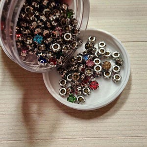 Peut inclure: Un récipient en plastique transparent déverse une collection de petites perles argentées avec des pierres précieuses colorées et à facettes. Les pierres précieuses sont de différentes couleurs, dont le rouge, le bleu, le vert et le rose. Les perles sont éparpillées sur un couvercle blanc et une surface en bois clair.