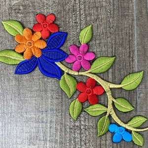 May include: Embroidered floral applique featuring a variety of colorful flowers and green leaves. The flowers are in shades of red, orange, pink, blue, and yellow. The leaves are green with yellow stitching. The applique is on a wooden surface.