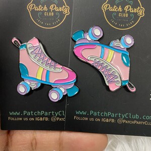 K&ouml;nnte beinhalten: Zwei rosa und blaue Rollschuh-Emaille-Pins mit wei&szlig;en Schn&uuml;rsenkeln und gelben Streifen. Die Pins befinden sich auf einem schwarzen Hintergrund mit dem Text "Patch Party Club" in einem goldenen Kreis.