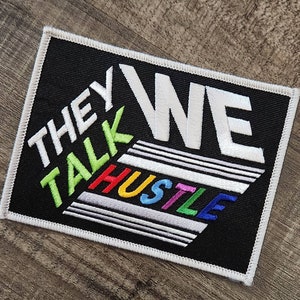 Pode incluir: Etiqueta bordada preta com texto branco que diz "They Talk We Hustle" em um esquema de cores arco-íris.