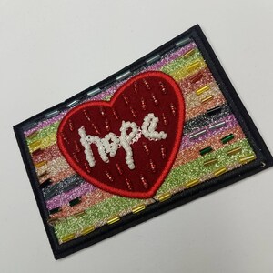 May include: A red heart-shaped patch with the word "Hope" spelled out in white beads. The heart is on a background of colourful stripes with glitter.