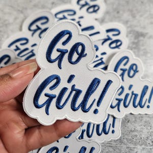 Pode incluir: Um remendo de tecido branco com texto bordado em azul que diz "Go Girl!" O remendo está sendo segurado em uma mão.