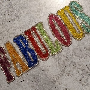 Può includere: La parola "FABULOUS" scritta in strass multicolori. Ogni lettera è di un colore diverso, a partire dal viola e terminando con il turchese.