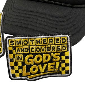 Puede incluir: Gorra de camionero negra con un parche amarillo y blanco brillante. El parche dice "SMOTHERED AND COVERED IN GOD'S LOVE!" en letras amarillas con un patrón de cuadros. La gorra tiene una visera curva y una parte trasera de malla.