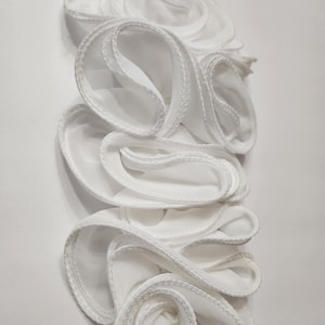 May include: A vertical arrangement of white fabric ruffles with a delicate, stitched edge. The ruffles are layered and create a flowing, floral-like design. The background is a plain white surface.