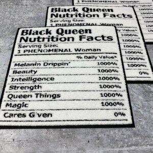 May include: Three rectangular white rugs with black borders. Each rug features the text "Black Queen Nutrition Facts," listing attributes like "Melanin Drippin'," "Beauty," and "Intelligence." The rugs are on a grey surface.