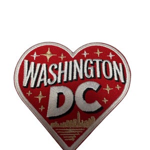 May include: A heart-shaped embroidered patch with a red background. The text WASHINGTON DC is stitched in white with a black outline. Gold-colored star accents are scattered across the surface, and a gold skyline silhouette sits at the bottom.