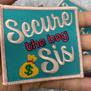 May include: A teal blue embroidered patch with pink stitching. The patch features a pink cursive font that says "Secure the bag sis" with a yellow money bag with a green dollar sign in the center.