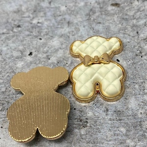 Op de afbeelding: Twee gouden teddybeer-bedels. Een bedel is effen goud, terwijl de andere wit is met een gestikt patroon en een gouden strik.