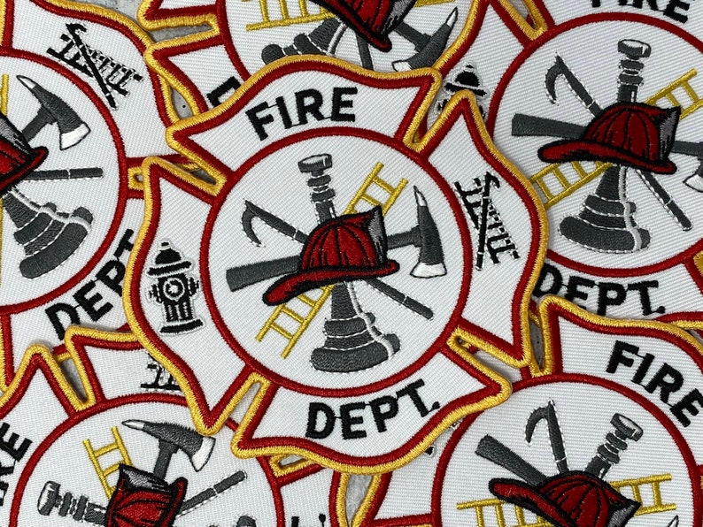 Op de afbeelding: Close-up van verschillende rode, witte en gouden brandweeremblemen met de tekst "FIRE DEPT." in het midden. De emblemen tonen een brandkraan, een ladder, een bijl en een brandweerhelm.