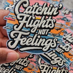 May include: Embroidered patch with the text "Catchin' Flights NOT Feelings" in white, outlined in black, on a blue, pink, and orange background. The patch features a stylized sunset with airplanes and clouds. The patch is held in a hand.
