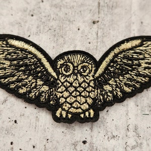 Könnte beinhalten: Eine goldene gestickte Eule mit schwarzen Flügeln, einem schwarzen Umriss und einem detaillierten Design. Die Eule fliegt mit ausgebreiteten Flügeln.