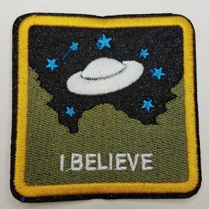 May include: A yellow square patch with a black background and a white flying saucer with the text "I BELIEVE" in white letters.