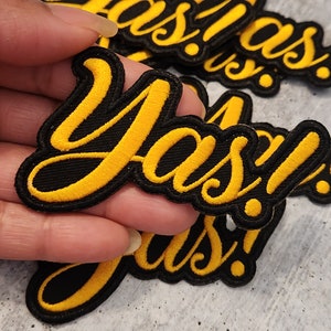 May include: A set of black and yellow iron-on patches with the word "Yas!" in a stylized font.
