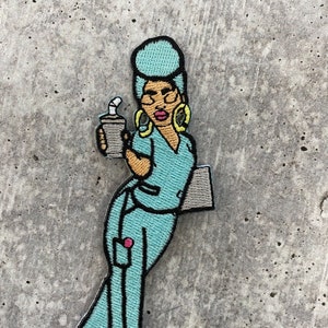 Könnte beinhalten: Ein gestickter Aufnäher mit einer Comic-Frau in einem hellblauen OP-Oberteil und einer Hose, die eine Kaffeetasse und eine Klemmbrett hält. Sie hat einen hellblauen Afro, große goldene Ohrringe und eine Brille.