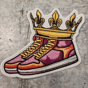 May include: Embroidered patch featuring a pair of sneakers with a crown. The sneakers are pink, orange, and purple with yellow laces. The crown is gold with fleur-de-lis details, outlined in white.