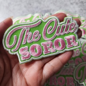 Puede incluir: Parche bordado verde y rosa con el texto "The Cute Soror" en una fuente cursiva.
