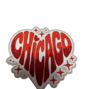 Op de afbeelding: Een geborduurde patch in de vorm van een hart met een witte achtergrond en een rood verlooptekst die CHICAGO spelt. Het ontwerp bevat kleine rode stervormige accenten en een witte rand, waarbij de rode letters naar beneden lijken te druipen.