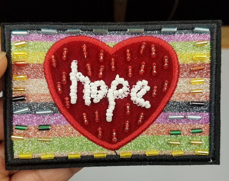May include: A red felt heart with the word "Hope" spelled out in white beads, surrounded by a colourful striped border. The heart is on a black background.