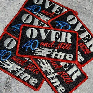 May include: A stack of black iron-on patches with red stitching. The patches feature white and blue embroidery that reads "Over 40 and still fine".