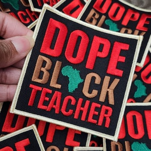 May include: A black square patch with white stitching around the edges. The patch has the words "Dope Black Teacher" embroidered in red and brown letters. A green outline of the continent of Africa is embroidered in the center of the patch.