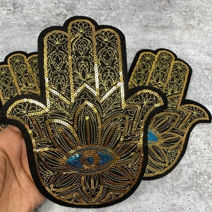May include: Two gold and black hamsa hand patches with a blue evil eye in the center. The patches are made of sequins and are embroidered with intricate designs.