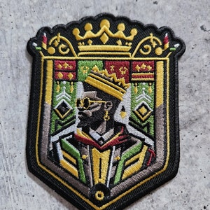 May include: A black and gold embroidered patch featuring a stylized image of a person wearing a crown, sunglasses, and a green and gold robe. The patch is shaped like a shield and has a gold crown at the top.