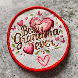 Puede incluir: Un parche blanco bordado con un borde rojo. El parche presenta un diseño de corazón con el texto "Best Grandma Ever" en letras doradas.