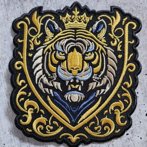 May include: A black and gold embroidered patch featuring a white tiger with a crown on its head. The tiger is surrounded by ornate gold scrollwork.