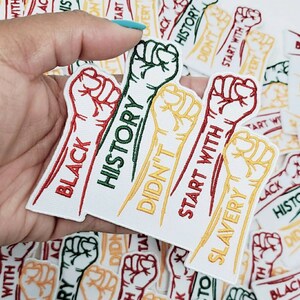 以下が含まれることがあります： 刺繍された拳とテキストが入った白い布パッチ。パッチには「Black History」、「Didn't Start With Slavery」、「Start With」などのフレーズが使用されています。拳は赤、緑、黄色です。