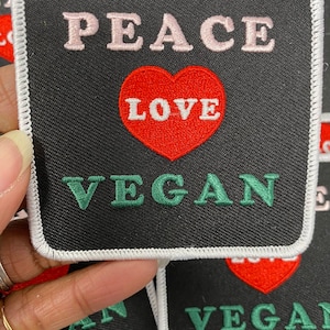 May include: A black fabric patch with white stitching around the edges. The patch has the words "Peace Love Vegan" embroidered on it in pink, red, and green thread.
