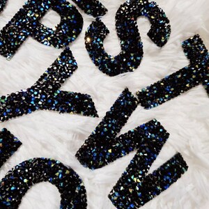 Peut inclure: Un ensemble de lettres en strass noires et bleues, probablement pour un projet d'artisanat ou pour personnaliser un article.