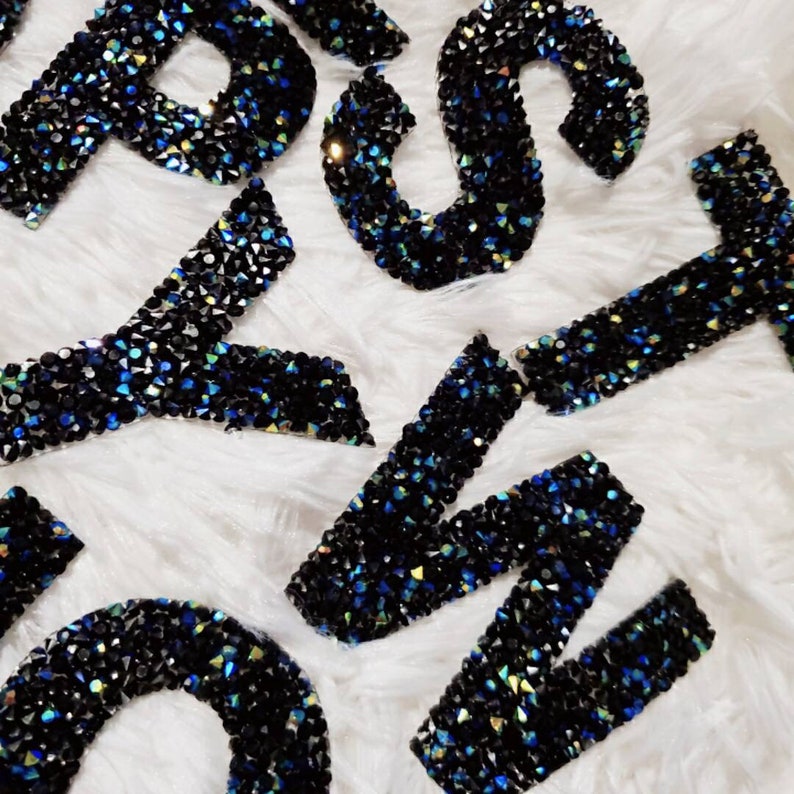Peut inclure: Un ensemble de lettres en strass noires et bleues, probablement pour un projet d'artisanat ou pour personnaliser un article.