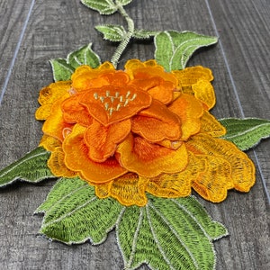 May include: Embroidered floral applique featuring a large orange and yellow flower with green leaves and stem. The flower has multiple layers of petals and detailed stitching. The leaves are a bright green with white stitching.