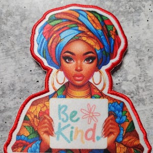 May include: A colorful embroidered patch featuring a woman with a patterned head wrap holding a sign that says "Be Kind." The sign has a flower drawn on it.