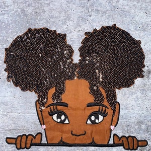 May include: Embroidered patch of a brown-skinned girl with large eyes and a smile. She has two large brown afro puffs with sequins. The girl is peeking over a brown banner.