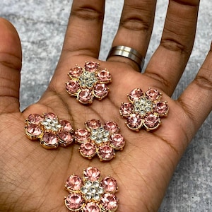 Könnte beinhalten: Mehrere blütenförmige Verzierungen mit rosa und klaren Kristallen. Die Blütenblätter sind rosa und die Mitte ist klar. Die Verzierungen haben eine goldfarbene Metallbasis. Die Artikel werden auf einer Hand präsentiert.