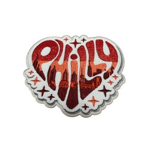 May include: An embroidered patch shaped like a heart with a white border. The interior features the word PHILY in a stylized font with a gradient of red, orange, and dark brown colors. The design includes a silhouette of a city skyline and stars.